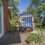 Terrasse mit Strandkorb, Tisch und Stühlen. Hintergrund: Grünfläche und Bäume.