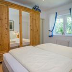 Schlafzimmer mit Doppelbett, Holzschrank und Fenster mit weißen Vorhängen.