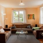 Living room with sofa, armchairs, and glass table. Windows with curtains. Wall art with flowers.