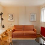 A room with an orange sofa, wooden table, chair, and two windows.