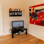 Living room with TV, wall art, and bookshelf. Wooden floor and door to kitchen.