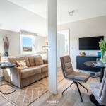 Living and dining area with sofa, TV, and table. Windows show landscape.