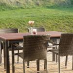 Holztisch mit vier Stühlen auf Terrasse. Hintergrund: grüner Rasenhang.