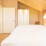 Schlafzimmer mit weißem Bett, Holzdecke und Tür zum Wohnbereich.