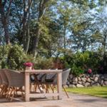 Holztisch mit Stühlen auf Terrasse, umgeben von Grün und Steinmauer.