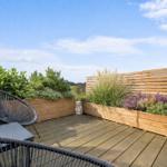 Terrasse mit Holzplanke, Pflanzenbeeten und Sitzmöbeln unter blauem Himmel.