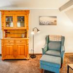 Ein Wohnzimmer mit Holzschrank, Sessel und Stehlampe. Küchenbereich ist sichtbar.