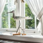 Zwei hölzerne Vogeldekorationen stehen auf dem Fensterbrett vor einem Fenster mit weißen Vorhängen.