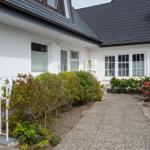 Weiße Hausfassade mit dunklem Dach, Eingangstür und Blumenbeeten entlang des Weges.