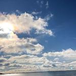 Sonnenlicht durchzieht den Himmel mit weißen Wolken über einem ruhigen Gewässer.