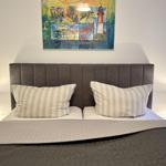 Doppelbett mit Kopfteil, Kissen und Decke. Wandbild mit Leuchte links und rechts.