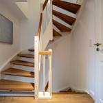 Treppenhaus mit Holztreppe und weißer Balustrade. Wandbild und Tür sind sichtbar.