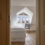 Schlafzimmer mit Dachschrägenfenster, weißem Bett und grauer Sitzgelegenheit.