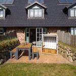 Haus mit Terrasse, Tisch, Bänken und Strandkorb. Holzwand und Dach mit Solarpanel.