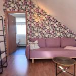 Ein Raum mit rosa Sofa, floraler Wandtapete und Holzboden. Eine Tür führt in einen anderen Raum.