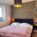 Schlafzimmer mit Bett, rosa Bettwäsche, dunkler Tapete und hängenden Pflanzen.