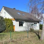 Ein zweistöckiges Haus mit grauer Fassade, Dachgauben und Garten mit Bäumen.
