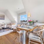 Living and dining area with sofa, table, and chairs. Wooden floor and large windows.
