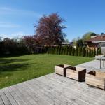 Holzterrasse mit Tisch und Kisten, grüner Garten mit Spielplatz und Bäumen.