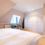 Bedroom with sloped ceilings, white duvet, two chairs, and white wardrobes.