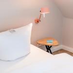 A bed with white linens, a pink wall lamp, and a small wooden table.