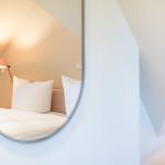 Through a round mirror, a bed with pillows and a lamp is visible.