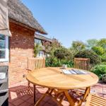 Terrace with wooden table and chairs in front of a house with thatched roof.