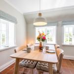Dining room with wooden table, chairs, and windows. Table set with flowers and bottle.