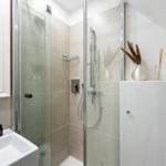 Dusche mit Glaswand, Waschbecken und Spiegel. Dekorative Vase und Duftstäbchen auf Schrank.