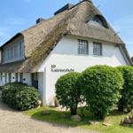 Haus mit strohgedecktem Dach und weißen Wänden, umgeben von grünen Sträuchern.