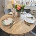 Runder Holztisch mit Platz für vier Personen, Blumenstrauß und Fensterblick.