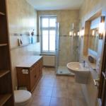 Badezimmer mit Dusche, Waschbecken und Toilette. Holzregal und Fenster sichtbar.