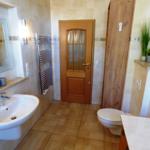 Badezimmer mit Waschbecken, Toilette und Heizung. Holztür und Pflanzen sind sichtbar.
