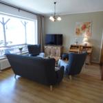 Wohnzimmer mit Sofa, Sesseln, Fernseher und Holzmöbeln. Große Fenster mit Blick auf Balkon.