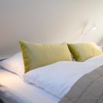 Ein Bett mit weißen Laken und zwei grünen Kissen. An der Wand hängen zwei weiße Stehlampen.