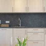 Küche mit weißen Schränken, grauer Rückwand und Arbeitsplatte. Tisch mit Blumen und Äpfeln.