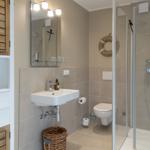 Badezimmer mit Dusche, Waschbecken und Toilette. Holzregal und Korb sind sichtbar.