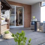 Terrasse mit Sitzgruppe, Glastür und Holzhaus. Pflanzen und Tisch sind sichtbar.