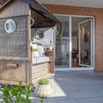 Holzkonstruktion mit Rattan, Sitzbank und Flasche im Freien. Hintergrund: Wohnbereich mit Glasfront.