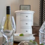 Tisch mit Weinglas, Flasche, Teller und Zitrusfrüchten. Hintergrund: weißer Schrank und Vorhang.