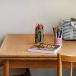 Holztisch mit Stuhl, Stifte und Zeichentrickbuch. Hintergrund: Wand und Koffer.