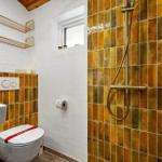 Bad mit Dusche, Toilette und Waschbecken. Wände mit goldenen Fliesen und Metallregalen.