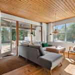 Wohnzimmer mit Sofa, Sessel und Holzboden. Große Fenster und Glastüren bieten Blick auf Garten.