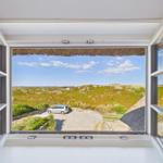 Durch das Fenster blickt man auf eine grüne Dünenlandschaft mit einem geparkten Auto.