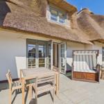 Terrasse mit Tisch, Stühlen und Strandkorb vor einem Haus mit Strohdach.