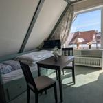 Room with bed, table and chairs, view onto balcony with sight on rooftops.