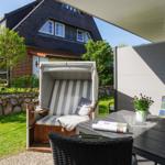 Terrasse mit Liege, Tisch und Stühlen. Hintergrund: Haus mit Steinmauer und Grün.
