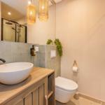 Badezimmer mit Waschbecken, Toilette und Dusche. Holzunterschrank und zwei Hängelampen.