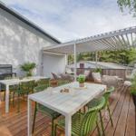 Terrasse mit Tischen, Stühlen und Pergola. Holzboden und Pflanzen sichtbar.