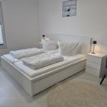 Schlafzimmer mit weißem Bett, Nachttischen und Stuhl. Fenster mit Jalousie und Wandbild.